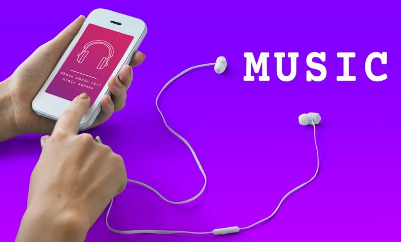 A person holding a white smartphone with headphones attached to it on a purple background